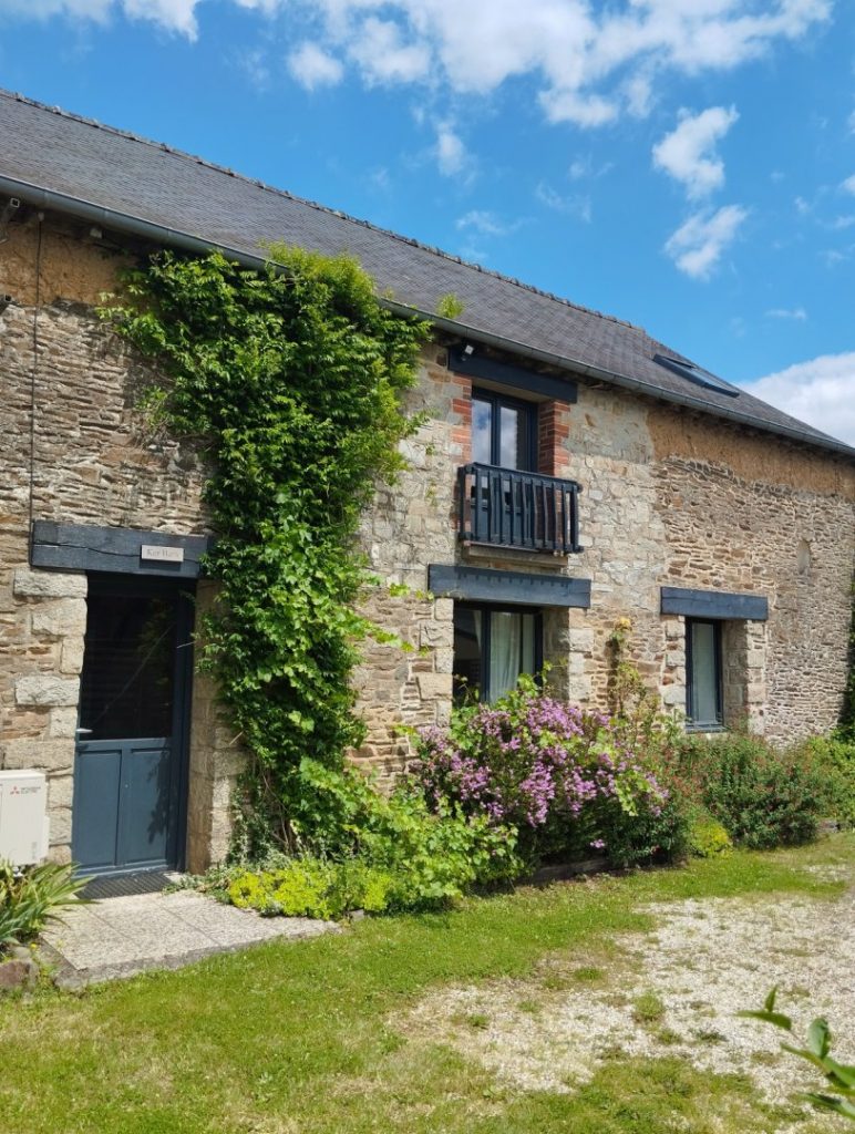 Vue de la cour pavée du gîte 15 personnes Ker Bara du Domaine de Beauregard en pierre, avec portail en bois donnant sur un jardin fleuri, situé en Bretagne.